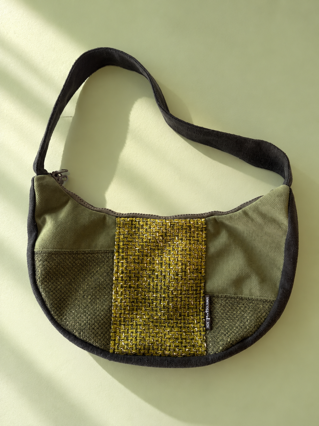 Olive-green patchwork crescent bag with a mustard-toned woven centre panel, dark edging and a dark shoulder strap.