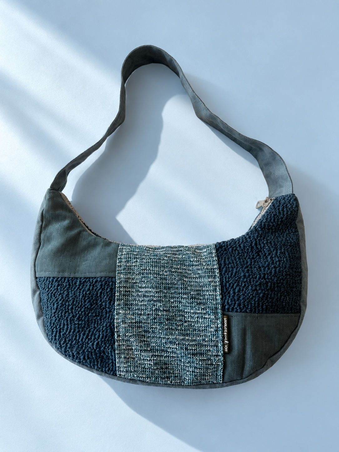 Teal patchwork crescent shoulder bag with a finely woven centre panel, deep teal raised-texture patches and a tonal strap.